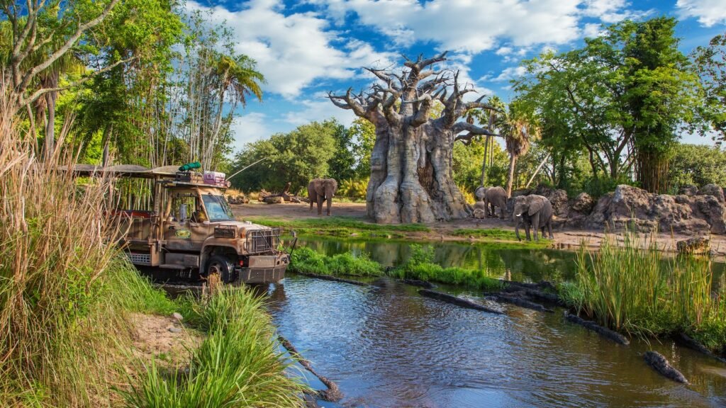 An open-air safari bus drives in a river past 3 elephants