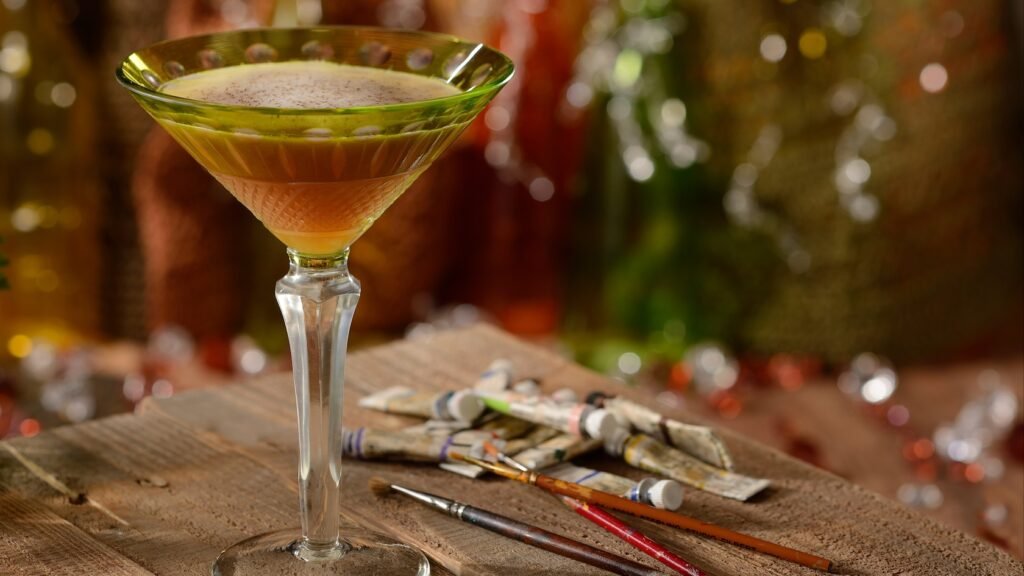 An elegant martini glass filled with a Night Monkey cocktail on a table beside tubes of watercolor paints and brushes